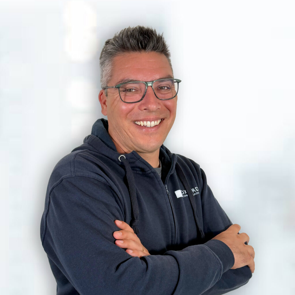 Porträt eines lächelnden Mannes mit Brille und verschränkten Armen vor hellem Hintergrund. Er trägt einen dunklen Kapuzenpullover mit Firmenlogo und blickt direkt in die Kamera.