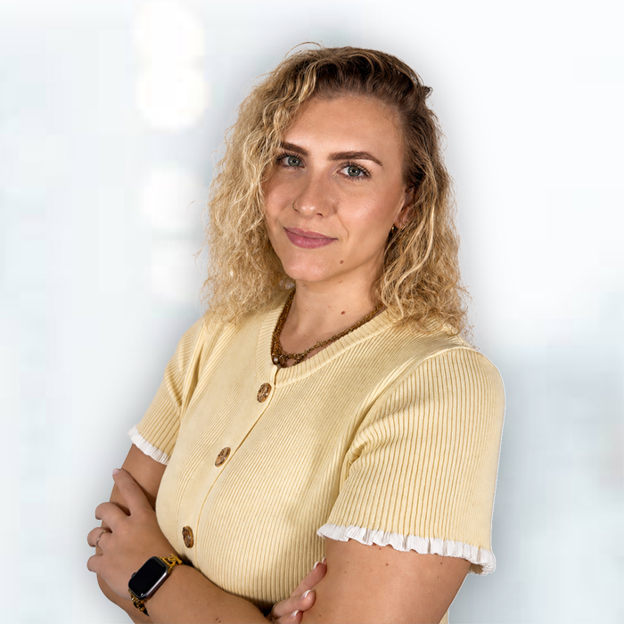 Porträt einer lächelnden Frau mit lockigen blonden Haaren und verschränkten Armen vor hellem Hintergrund. Sie trägt ein gelbes Oberteil mit Knopfleiste, Schmuck sowie eine Smartwatch und blickt direkt in die Kamera.